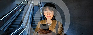 Happy smiling young woman, standing on escalator, going down, holding smartphone in both hands, chatting on mobile phone