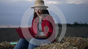 Happy smiling young woman in straw hat surfing Internet on laptop sitting in twilight on hay bale. Portrait of cheerful