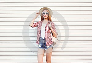 Happy smiling young woman in summer round straw hat, checkered shirt, shorts posing on white wall