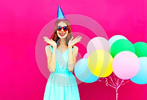 Happy smiling young woman is surprised in a birthday cap with an air colorful balloons over pink