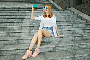 Happy smiling young woman taking selfie picture by smartphone sitting on the stairs on city street