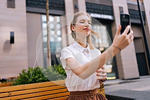 Happy smiling young woman taking selfie using smartphone and holding cup with takeaway coffee in summer day sitting on