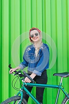 Happy smiling young woman wearing sunglasses and a denim jacket stands with a green bike