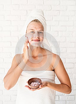 Happy smiling young woman in white bath towels applying scrub on her face and neck