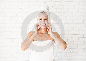 Happy smiling young woman in white bath towels applying scrub on her face and neck