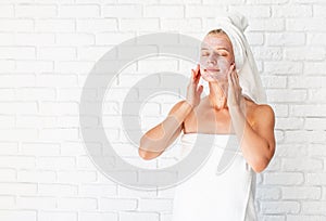 Happy smiling young woman in white bath towels applying scrub on her face and neck