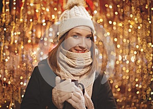 Happy smiling young woman in winter clothes holding cup with christmas lights in background