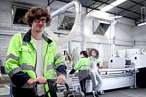 Happy smiling young worker man with curly hair wearing glasses and safety vest working in factory, industrial male engineer