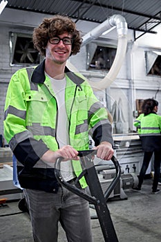 Happy smiling young worker man with curly hair wearing glasses and safety vest working in factory, industrial male engineer