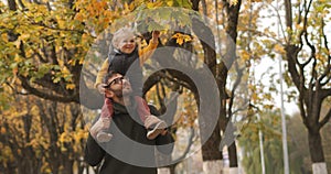 Happy son and father are walking in forest at autumn, baby is sitting on shoulders of dad and viewing leaves of tree