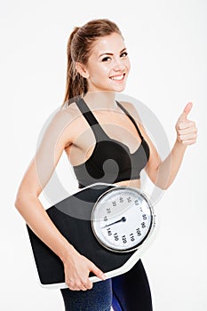 Happy sportswoman holding weight scales and showing thumbs up gesture