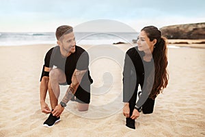 Happy sporty man and woman couple wearing sportswear and tying shoes, preparing for working out by seaside
