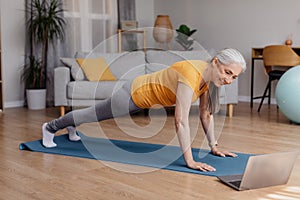Happy sporty senior woman standing in plank on yoga mat, working out to online sports video at home