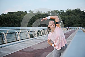 Happy sporty woman smiles toothy smile, exercising outdoors on the bridge, focusing on stretching during morning workout at