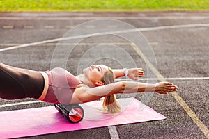 Happy sporty woman training on mat outdoor summer day, using foam roller massager on shoulder and back for relaxation