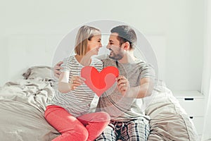 Happy strong marriage. Happy funny couple in love holding red paper heart. Heterosexual family man and woman sitting on bed in