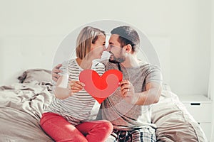 Happy strong marriage. Happy funny couple in love holding red paper heart. Heterosexual family man and woman sitting on bed in
