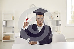 Happy student in black graduation robe and hat sitting at desk and showing his diploma