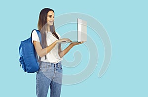 Happy student girl with backpack standing on blue background and working on laptop PC