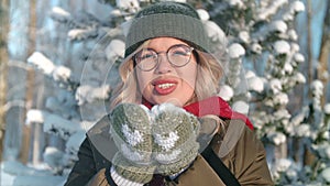 Happy student girl blowing out snow from palms in forest in park, portrait of woman in winter day