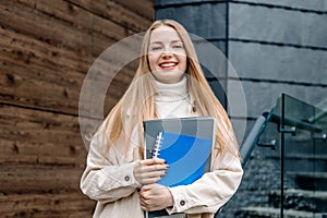 Happy student girl holds folders notebooks books in hands smiles against the background of a modern university building