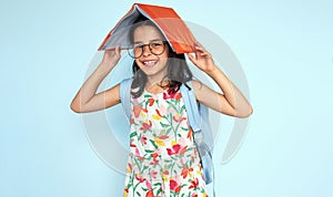 Happy student girl putting and holding academic book on her head and looking at the camera, wearing colorful dress, eyeglasses,