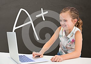 Happy student girl at table using a computer against grey blackboard with A+ text