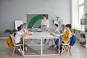 Happy students sitting around table in classroom, reading books and talking with teacher