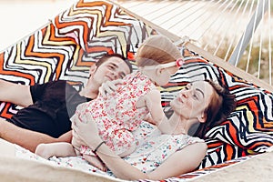 Happy stylish family with cute daughter relaxing in hammock on summer vacation in evening sun light on the beach. hipster couple