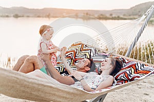 Happy stylish family with cute daughter relaxing in hammock on summer vacation in evening sun light on the beach. hipster couple