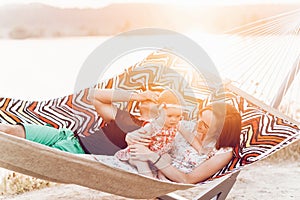 Happy stylish family playing with cute daughter, relaxing in hammock on summer vacation in evening sun light on the beach. hipster