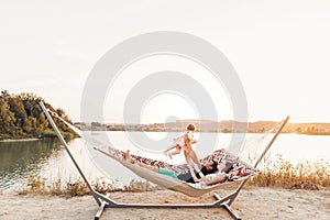 Happy stylish family playing with cute daughter, relaxing in hammock on summer vacation in evening sun light on the beach. hipster