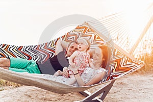 Happy stylish family playing with cute daughter, relaxing in hammock on summer vacation in evening sun light on the beach. hipster