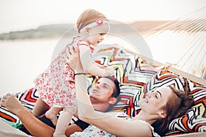 Happy stylish family relaxing in hammock on summer vacation in evening sun light on the beach. hipster couple with child resting