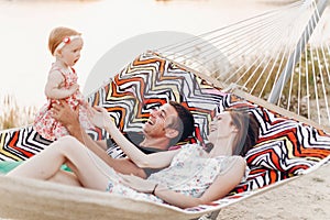 Happy stylish family relaxing in hammock on summer vacation in evening sun light on the beach. hipster couple with child resting