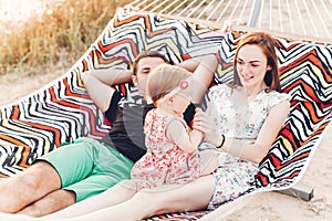 Happy stylish family relaxing in hammock on summer vacation in evening sun light on the beach. hipster couple with child resting