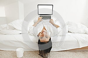 Happy stylish girl  lying on bed with laptop with blank screen and smiling in white modern room. Young cheerful woman shopping