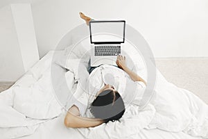 Happy stylish girl lying on bed with laptop with blank screen in white modern room. Young woman looking at empty display, shopping