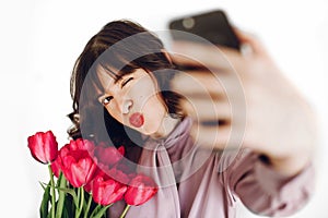 Happy stylish girl taking selfie on phone and holding pink tulip