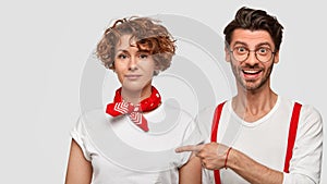 Happy stylish man with trendy haircut, stands near curly woman, wear white clothes, point at free space, advertise