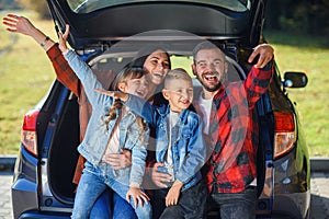 Happy stylish parents with their cute lovely children are making funny selfie on smart phone while sitting in the trunk