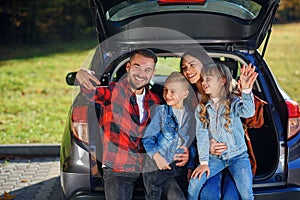 Happy stylish parents with their cute lovely children are making funny selfie on smart phone while sitting in the trunk