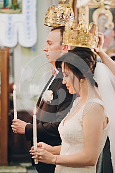 Happy stylish wedding couple holding candles with light under golden crowns during holy matrimony in church. Bride and groom