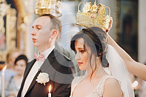 Happy stylish wedding couple holding candles with light under golden crowns during holy matrimony in church. Bride and groom