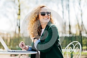 Happy stylish woman with curly light hair wearing sunglasses working with laptop outside in park typing necessary documents turnin