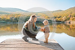 Happy stylish woman holding newborn baby over lake outdoor in sun light.