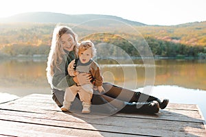 Happy stylish woman holding newborn baby over lake outdoor in sun light.