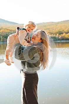 Happy stylish woman holding newborn baby over lake outdoor in sun light.
