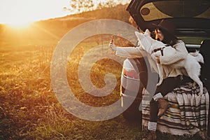 Happy stylish woman taking selfie photo with cute dog in car trunk in sunset light in field. Young hipster female using phone with
