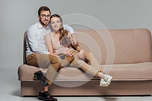 Happy stylish young couple with smartphone smiling at camera while sitting together on sofa